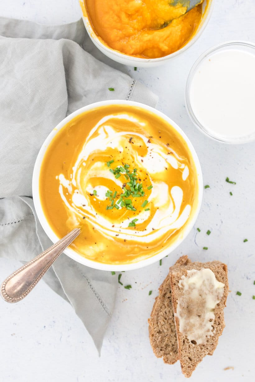 pumpkin soup with coconut milk swirled on top, in a white bowl with pumpkin and bread on the side