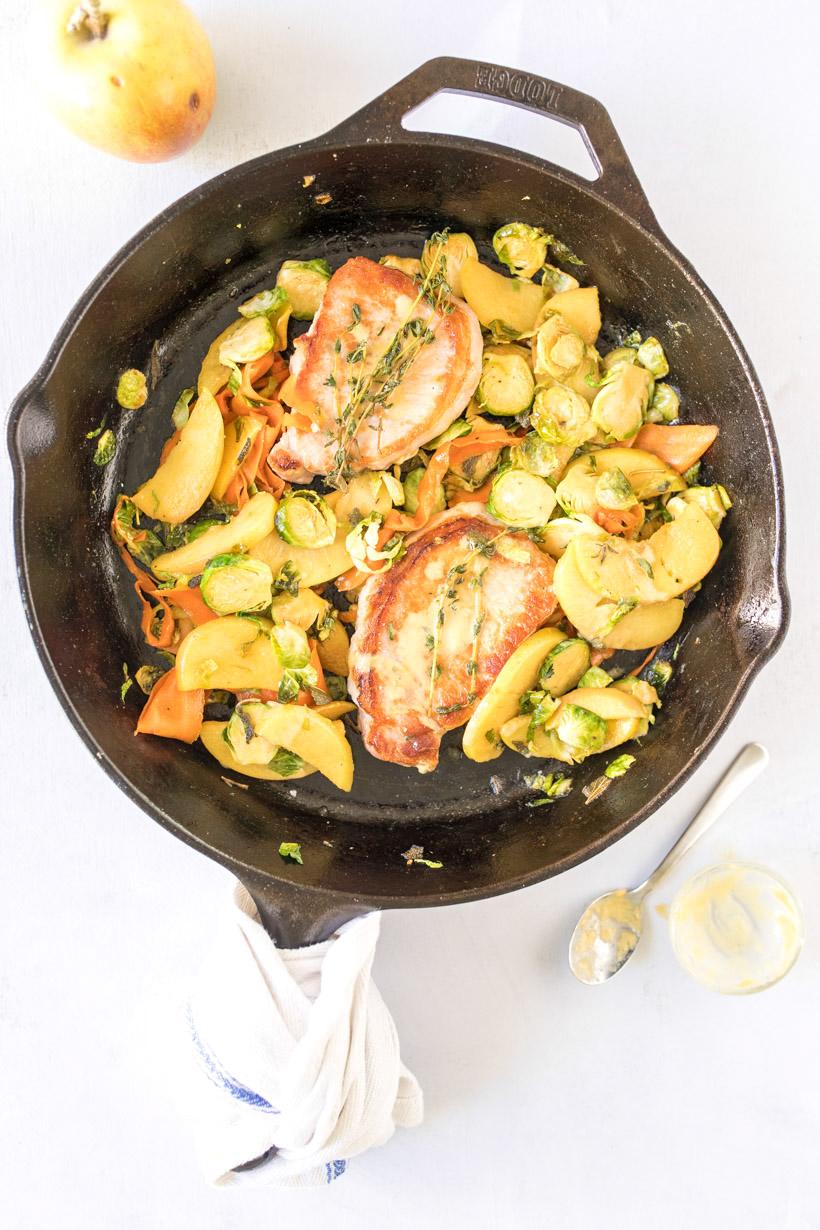 two pork chops, apples, carrots, and brussels sprouts in a cast iron skillet