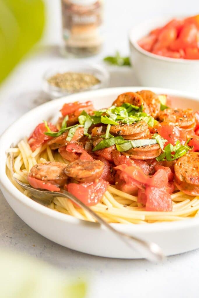simple pasta in a white bowl with sausage on top and ingredients behind