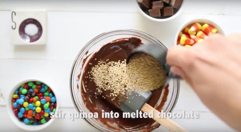 a hand pouring toasted quinoa into melted chocolate in a glass bowl with candies surrounding it