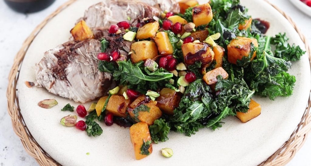 closeup of salad with kale, butternut squash, chopped pistachios, and pomegranate aerils with pork loin on a plate
