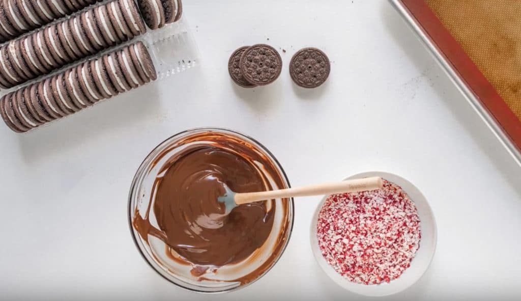 a bowl of melted chocolate, a bowl of crushed peppermint candies and oreos on a table