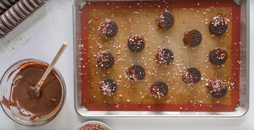 13 chocolate dipped oreos with crushed peppermint sprinkled on top on a baking sheet next to a bowl of melted chocolate
